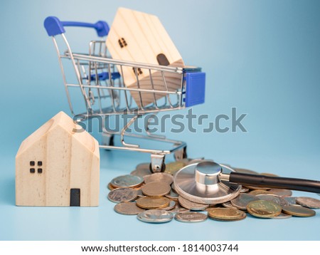 A model wooden house on a shopping cart With a pile of coins and stethoscope On a blue background. Mortgage concept. Money and house. Financial status.