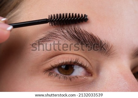 Modeling brows, gorgeous charming lady with beauty face, healthy clear skin, holds brush in hand and paint her eyebrows. Macro close up of brows.