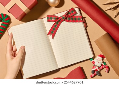 The model's hand opens a blank notebook with a red bow on top, surrounded by red and brown scrolls and a few other Christmas decorations. A close-up of the Christmas model from above. - Powered by Shutterstock