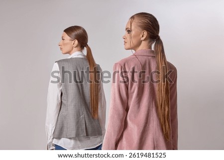 Models pose back-to-back in a studio, dressed in stylish neutral outfits with long hair pulled back. focus is on contemporary fashion and the unique textures of their clothing choices.