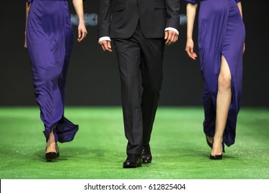 Models Walk The Runway During A Fashion Show. Fashion Catwalk Event Showing New Collection Of Clothes. Two Women In Blue Dresses And A Man In The Middle. Green Stage.