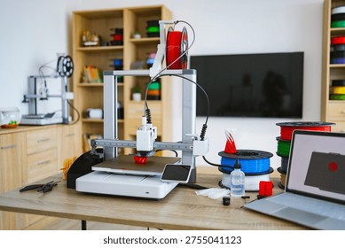 A modern 3D printer is actively creating a red object on its heated bed, surrounded by spools of filament and tools on a wooden desk