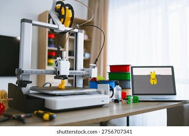 A modern 3D printer is actively creating a small yellow object on its platform, with spools of filament and a laptop nearby