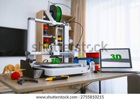 A modern 3D printer is actively creating green plastic letters on its build plate while a laptop displays the word 3D