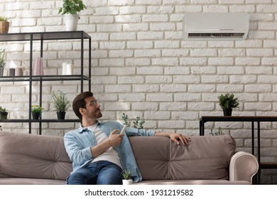 Modern Ac Is My Choice. Happy Male Owner Of New Apartment Sit On Couch At Home Hold Remote Controller Set Comfy Temperature On Air Conditioner. Young Man Control Climate Indoors Enjoy Fresh Cool Air