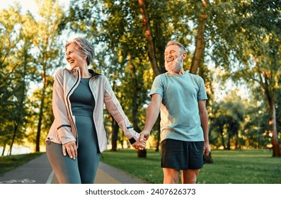 Modern active cheerful sports couple of senior people holding hands and talking cheerfully outdoors together. The woman turned her face to the rays of the sun and laughs. The joy of life. - Powered by Shutterstock