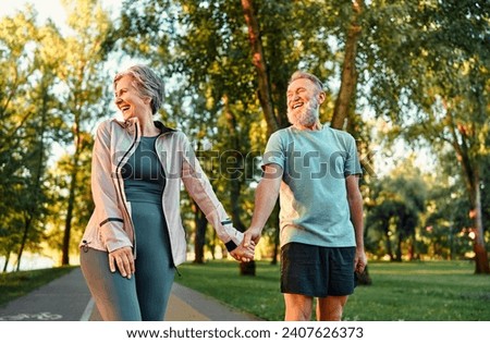 Modern active cheerful sports couple of senior people holding hands and talking cheerfully outdoors together. The woman turned her face to the rays of the sun and laughs. The joy of life.