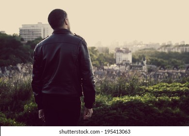 Modern Adult Sad Man Turned Back. Man On The Background Of The City And Nature At Sunset. A Man In A Black Jacket S Skin Looks And Thinks About Something.