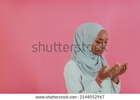 Modern African Muslim woman makes traditional prayer to God, keeps hands in praying gesture, wears traditional white clothes, has serious facial expression, isolated over plastic pink background