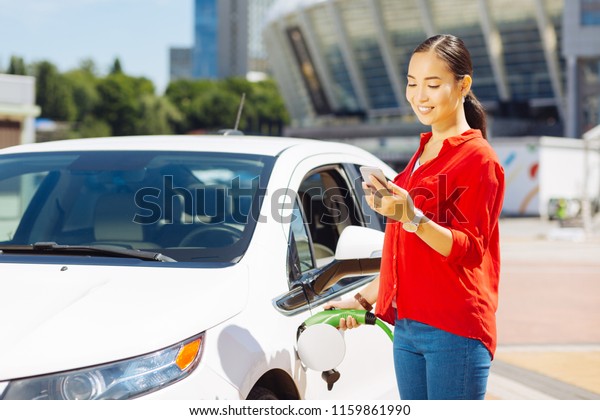 Modern age. Confident young woman looking\
at her smartphone while holding a fuel\
nozzle