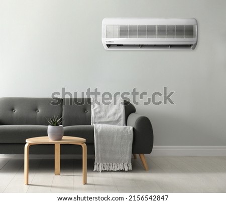 Modern air conditioner on white wall in room with stylish grey sofa