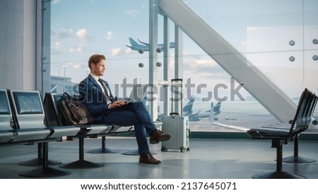 Modern Airport Terminal: Handsome Businessman Working on Laptop Computer While Waiting for His Flight. Man Sitting in a Boarding Lounge of Big Airline Hub with Airplanes Departing and Arriving