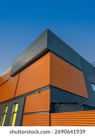 Modern angular building facade featuring vibrant orange and sleek gray metal panels, set against a clear blue sky, highlighting contemporary urban architecture.
