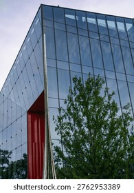 Modern angular commercial building featuring a large reflective glass facade and a striking red recessed entrance, framed by a green tree. The scene represents contemporary corporate headquarters, bold urban design, and professional business environments.