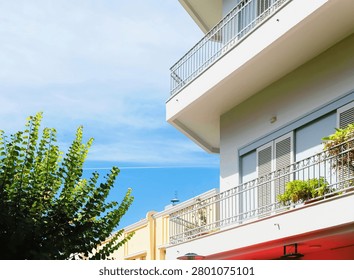 Modern apartment building presenting multiple balconies with metal railings, green potted plants, and clear blue sky background