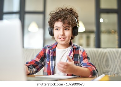 Modern approach to learning. Portrait of joyful latin american little boy wearing headphones, looking at the screen of a laptop while having online lesson at home - Powered by Shutterstock