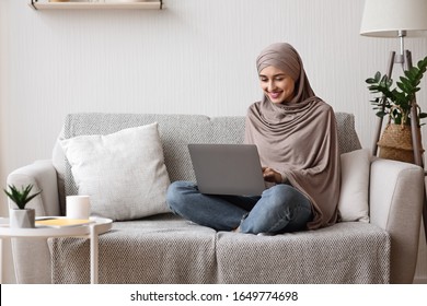 Modern Arab Girl In Hijab Using Laptop At Home, Working Remotely While Sitting On Couch In Living Room