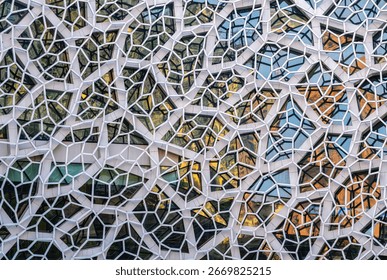 a modern architectural facade featuring a complex, geometric pattern of white, hexagonal and irregularly-shaped frames. The frames enclose various glass panels, creating a mosaic-like effect.