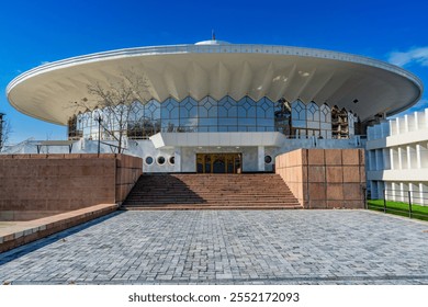 A modern architectural marvel featuring a circular design with a unique domed roof and geometric glass windows. The structure is complemented by wide steps, a tiled pathway, and clear blue skies. - Powered by Shutterstock