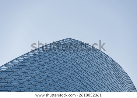 A modern architectural structure features a sleek, curved surface with grid of triangular glass panels. The sky provides a clear backdrop, emphasizing building's futuristic design.