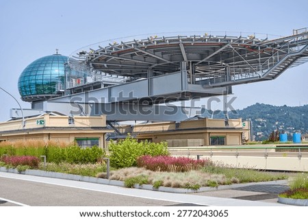 Modern architectural structure featuring the Pinacoteca Giovanni e Marella Agnelli atop the former Fiat Lingotto factory in Turin, Italy. The image prominently displays Renzo Piano's distinctive glass dome nicknamed 'La Bolla' (The Bubble) with its blue-tinted transparent panels, alongside a dramatic cantilevered steel helipad and testing track structure extending over the building edge. Below the metallic infrastructure sits the beige-brown historic industrial building with visible exit signs and ventilation equipment. The foreground features landscaped roof gardens with ornamental grasses, flowering shrubs, and paved walkways. In the background, tree-covered hills rise under a clear pale blue sky, indicating a sunny day. The composition showcases the contrast between industrial heritage and contemporary architectural intervention.