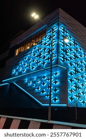 Uma estrutura arquitetônica moderna iluminada com luzes LED azuis impressionantes à noite. A fachada apresenta intrincados padrões e formas geométricas, criando um olhar futurista e artístico. 