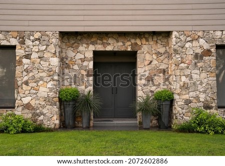 Modern architecture. Residential facade design. Closeup view of new house iron front door, stonewall and decorative plants Buxus sempervirens, also known as boxwood bush, growing in pots.