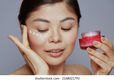 Modern Asian Female With Facial Cream Jar And Eye Cream On Face Against Blue Background.