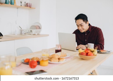 Modern Asian Man Working On Laptop In The Kitchen At Breakfast.