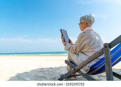 Modern Asian senior businesswoman working on digital tablet with internet at tropical beach in sunny day. Elderly retired woman enjoy outdoor lifestyle with using technology on summer holiday vacation - Powered by Shutterstock