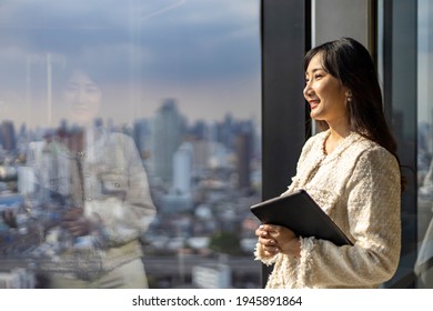 Modern asian woman entrepreneur holding digital tablet looking out the window glass with skyscraper and cityscape on the background with copy space - Powered by Shutterstock