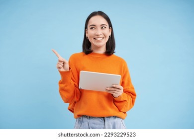 Modern asian woman with tablet, pointing at empty space for promo offer, banner, smiling and looking at advertisement, blue background. - Powered by Shutterstock