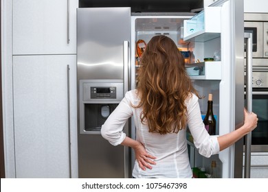 Modern attractive woman with long hair standing in the kitchen opening the frige door and looking inside.