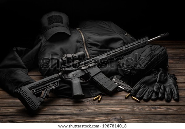 Modern automatic rifle with a telescopic sight\
on a dark background. Jacket, cap, gloves on a wooden table. The\
uniform of a guard or a\
mercenary.