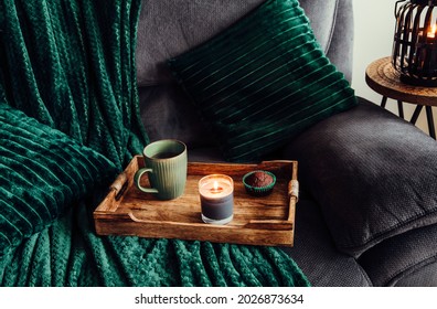 Modern autumn hygge set in living room. Dark green interior elements, soft pillows, plaid on sofa with chocolate muffin, aroma drink mug on wood tray and scented candle burning.  - Powered by Shutterstock