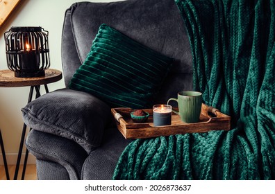 Modern Autumn Hygge Set In Living Room. Dark Green Interior Elements, Soft Pillows, Plaid On Sofa With Chocolate Muffin, Aroma Drink Mug On Wood Tray And Scented Candle Burning. Hygge Home.