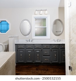 A modern bathroom featuring a dual sink vanity, decorative mirrors, wood flooring, and natural light from a window. - Powered by Shutterstock