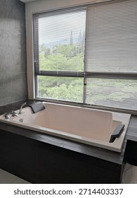 A modern bathroom featuring a white rectangular built-in bathtub set against a large window with horizontal blinds, offering a scenic outdoor view. The tub is framed by dark charcoal 