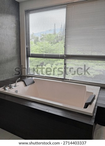 A modern bathroom featuring a white rectangular built-in bathtub set against a large window with horizontal blinds, offering a scenic outdoor view. The tub is framed by dark charcoal 