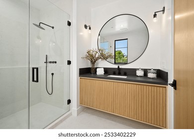 Modern bathroom with glass shower, round mirror, wooden vanity, and natural light from a window. - Powered by Shutterstock