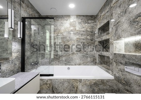 A modern bathroom with grey marble-patterned walls, white bathtub, glass shower screen with black frame, built-in niches, speckled countertop and sleek pendant lights, luxurious and minimalist.