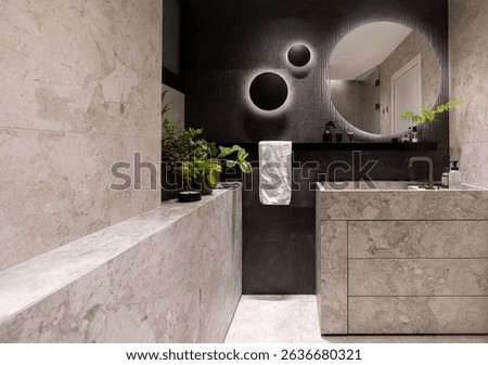 Modern bathroom interior featuring light gray natural stone tiles, matte black accents, a round backlit mirror, built-in stone vanity, and green plants adding warmth 