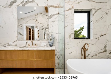 Modern bathroom interior with marble walls, wooden vanity, and gold fixtures, featuring a large mirror and a window with a view of greenery. - Powered by Shutterstock