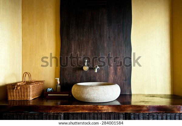 Modern bathroom interior with original white stone sink,\
clock, wooden baskets and soap dispenser. Wooden interior of spa in\
yellow and gold colors. Selective focus, only part of bathroom is\
in focus 