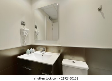 Modern Bathroom With White Porcelain Sink With Dark Wood Cabinet With Frameless Mirror On The Wall, Hairdryer With Plug And Shiny Tiles