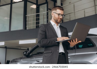 Modern bearded man in glasses and suit vehicle sales consultant using laptop inside car dealership - Powered by Shutterstock