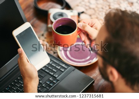 Modern bearded man using cellphone and drinking coffee with laptop.