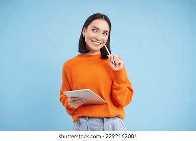 Modern beautiful asian woman with digital tablet and pencil, taking notes, writing on her gadget, doing homework, working, standing over blue background. - Shutterstock ID 2292162001