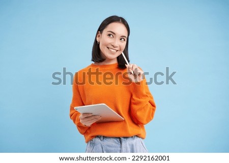 Modern beautiful asian woman with digital tablet and pencil, taking notes, writing on her gadget, doing homework, working, standing over blue background.