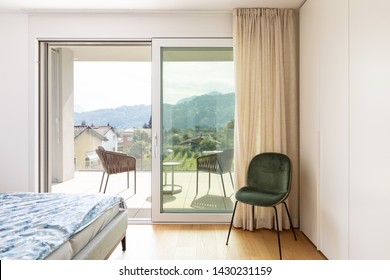 Modern bedroom with bed and velvet chair. Window overlooking nature. Nobody inside. 